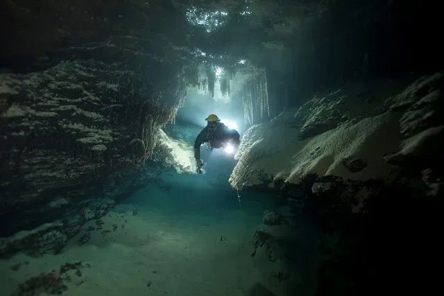 A cave diver in a helmet swimming towards the camera through a decorated passage, lit from behind, a thin guideline running along the floor