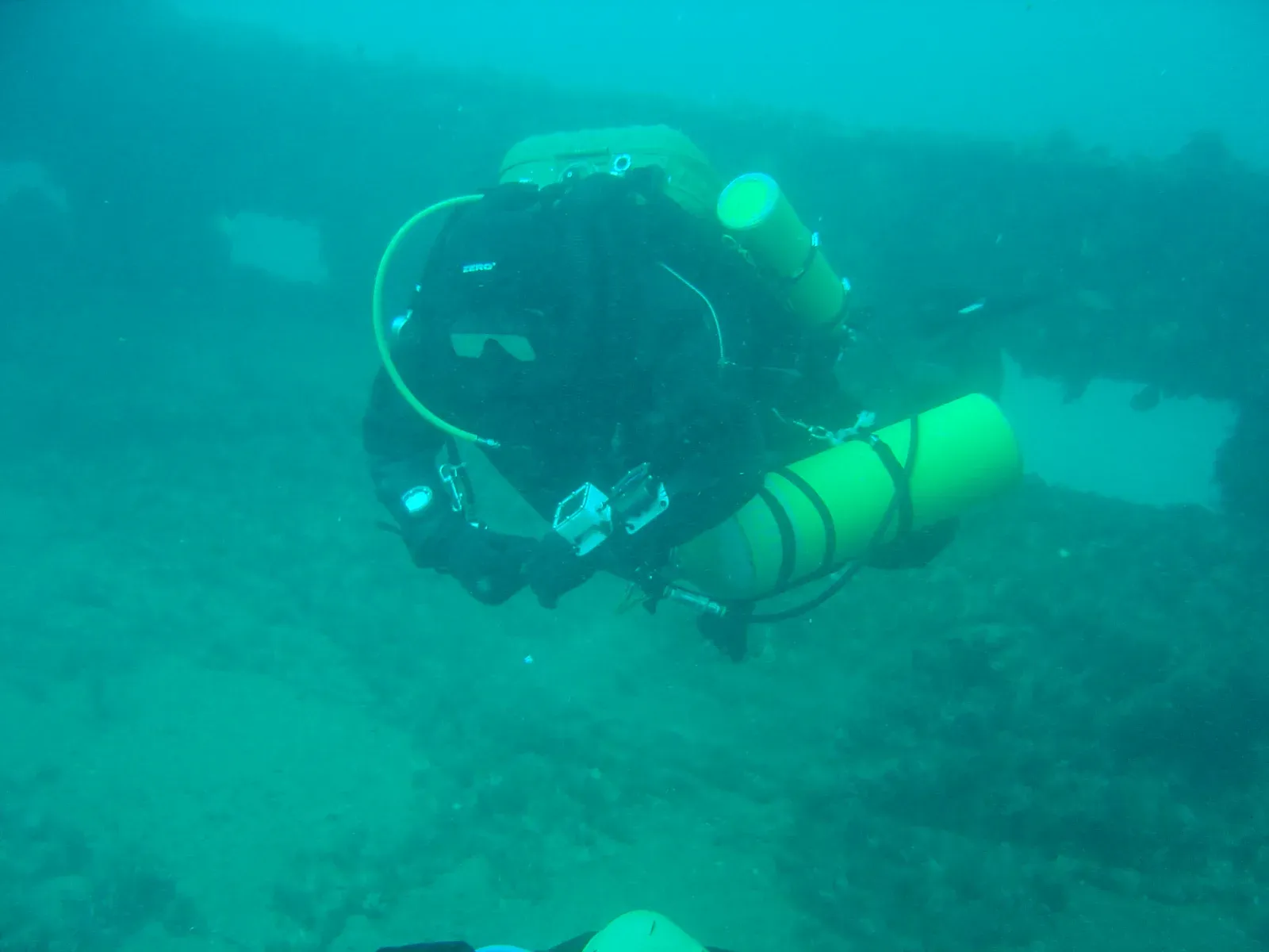 A rebreather diver hovering over a wreck in green water, carrying two yellow bailout cylinders clipped alongside the unit, releasing no bubbles