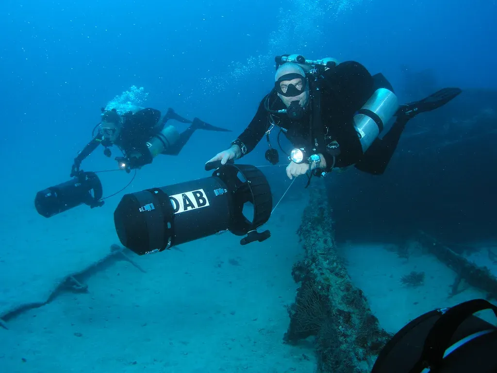 Two divers in twin cylinders riding cylindrical scooters low over a wreck in clear blue water