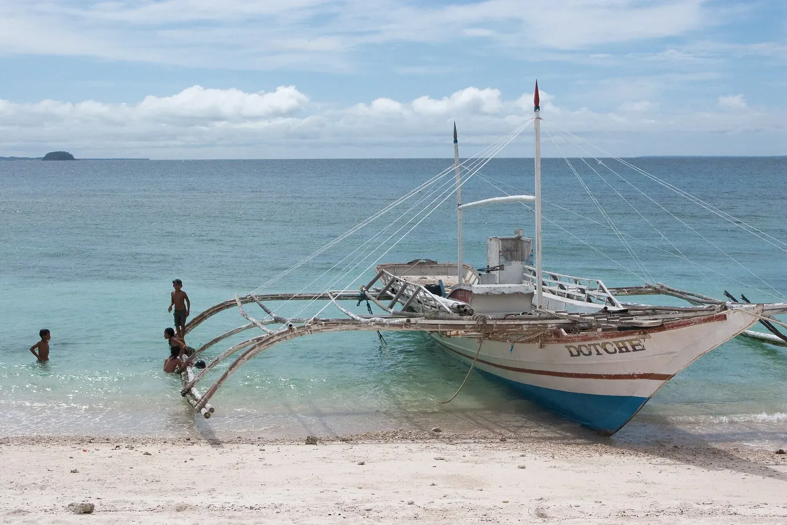 A white outrigger bangka boat beached on white sand at Malapascua, Philippines, with swimmers beside it
