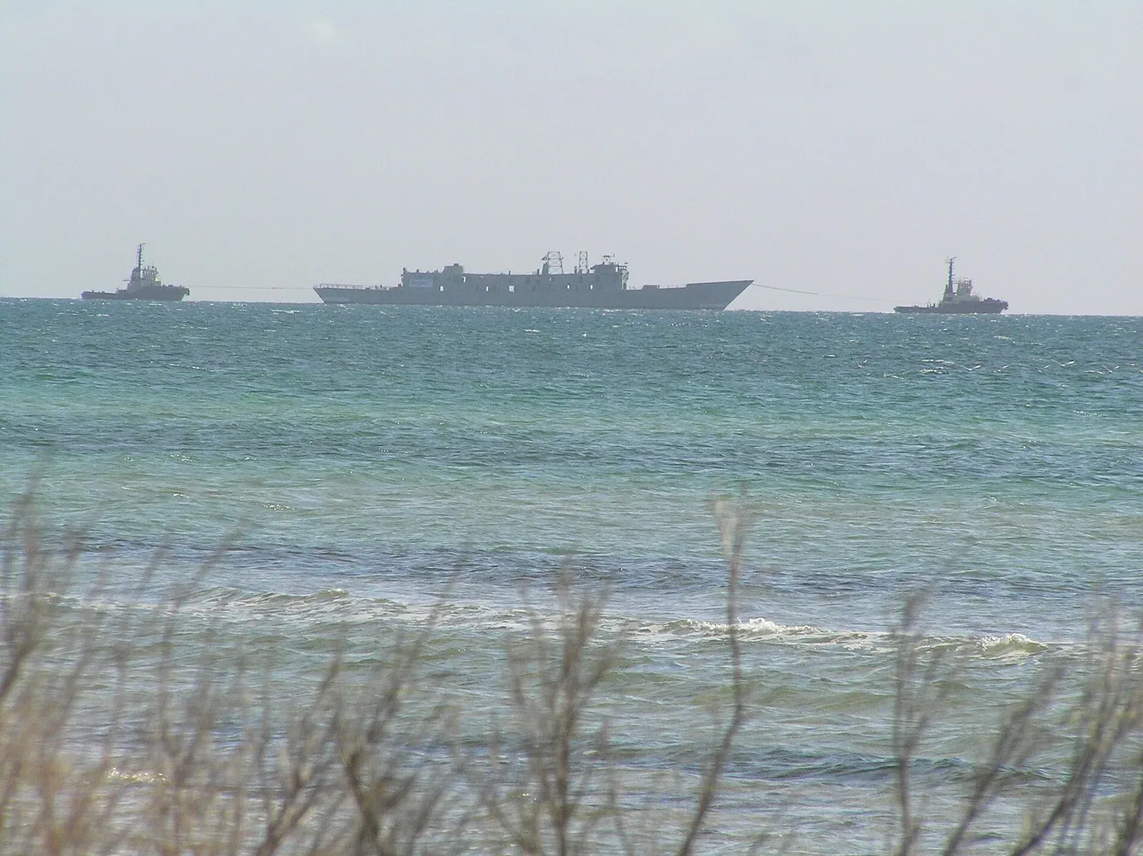 A grey warship on the horizon under tow between two tugs, seen from a beach across choppy green water