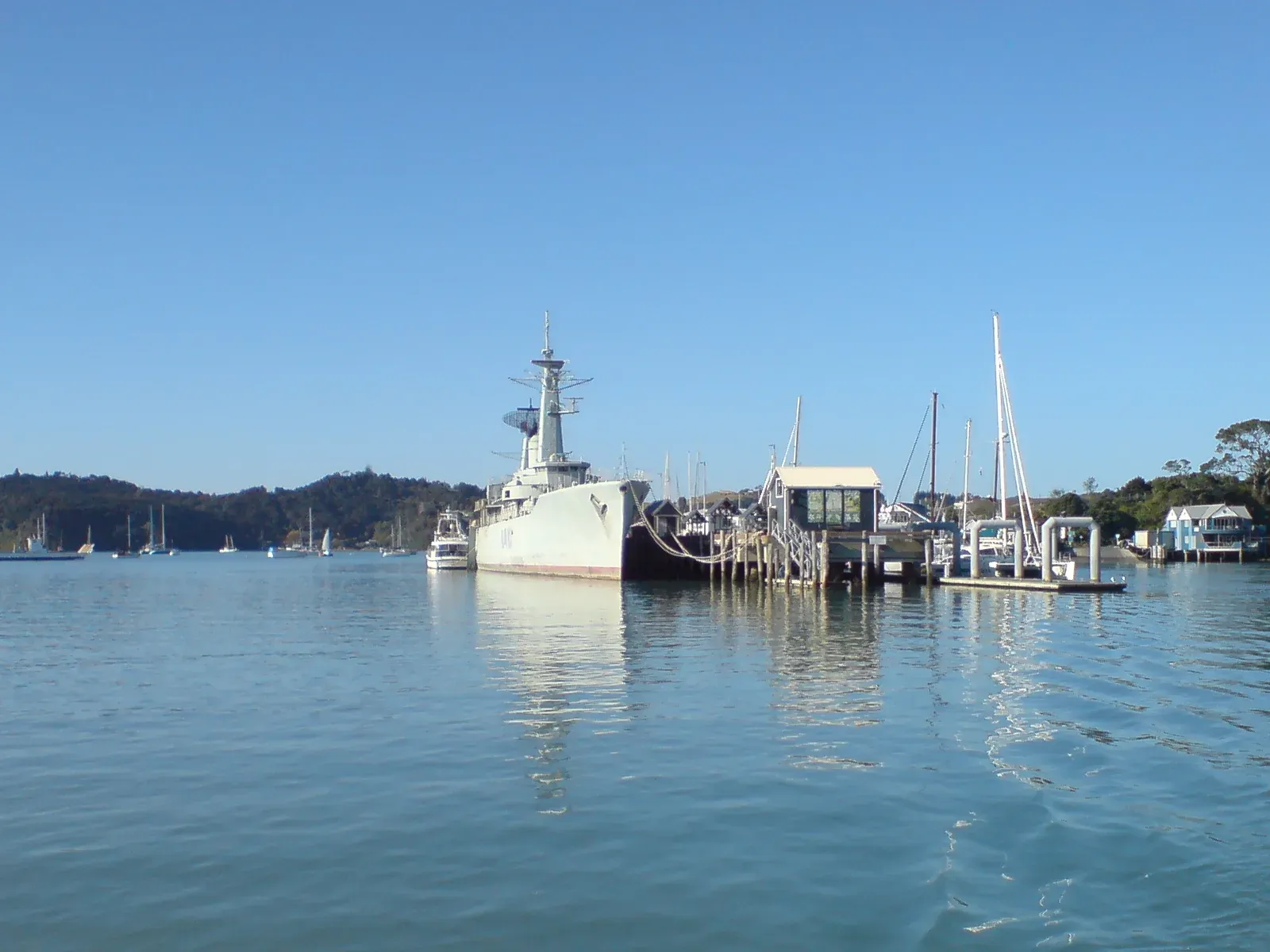 A grey navy frigate moored at a small wooden quay among yachts on a calm, sunny harbour