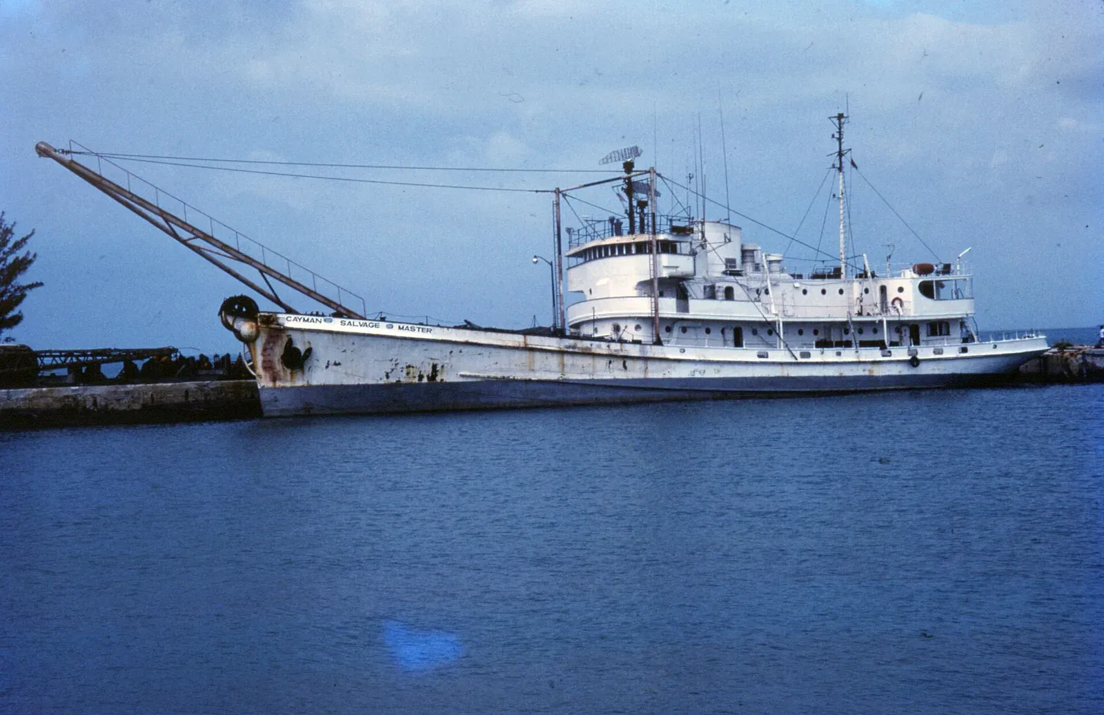 A white salvage ship with a long boom crane and tall mast moored alongside a stone harbour wall