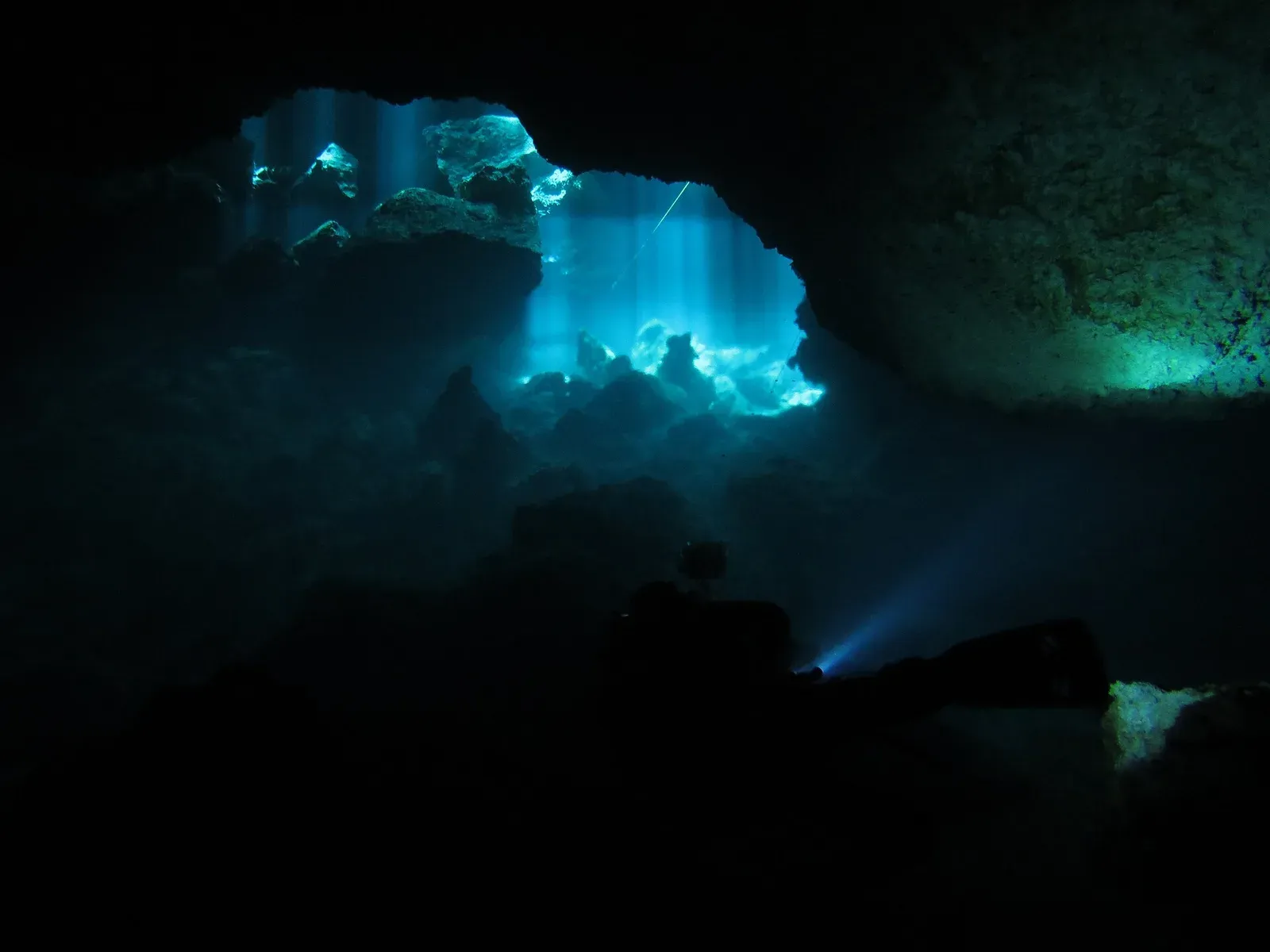 Sunbeams cutting through the dark chambers of Cenote Chac Mool, with a diver's torch beam glowing through hazy layered water