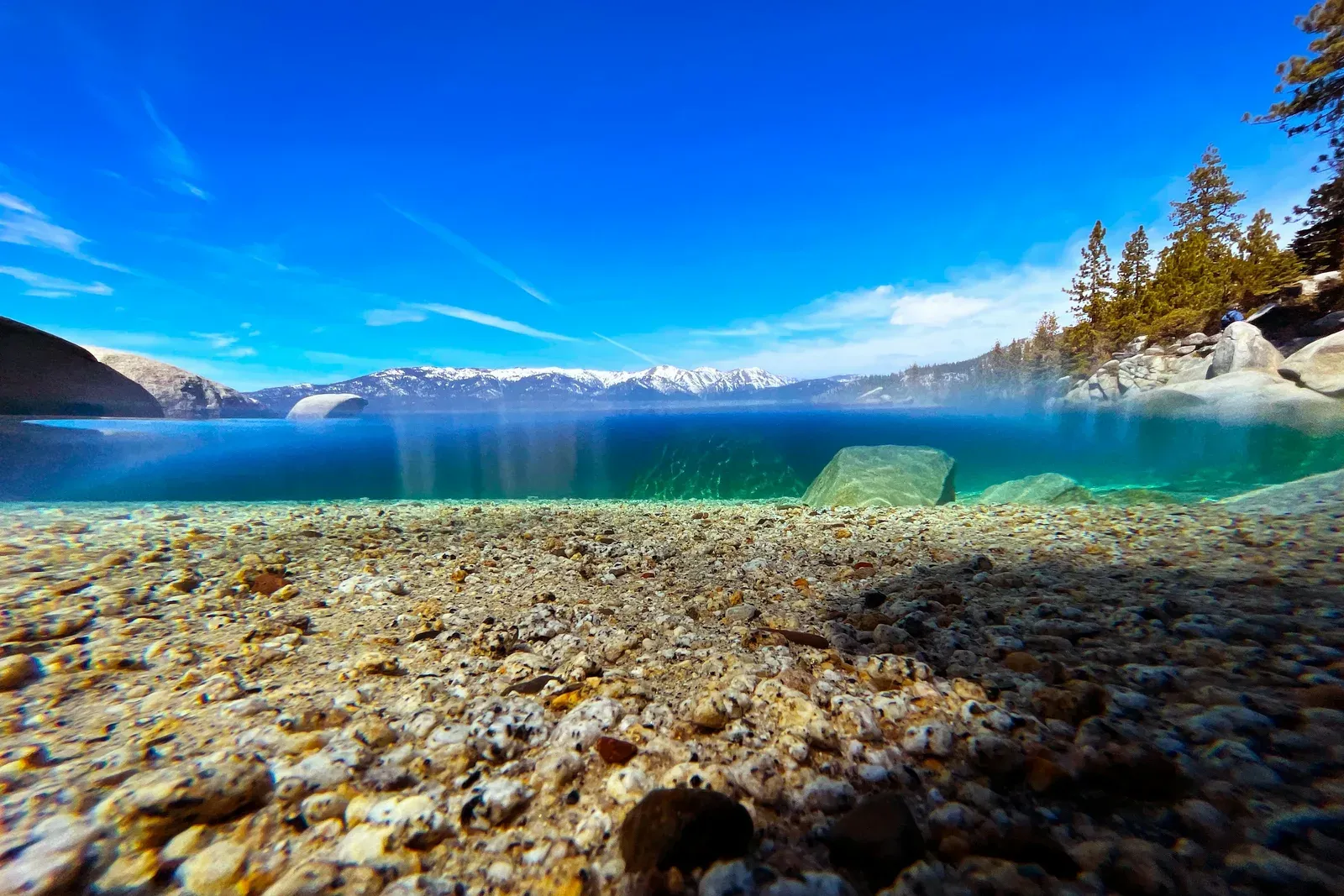 Split-level view of Lake Tahoe with snow-capped mountains above the surface and granite pebbles and boulders below