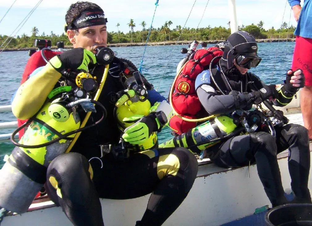 Two technical divers kitting up on a boat gunwale, slung stage cylinders with gas-content labels clipped to their harnesses