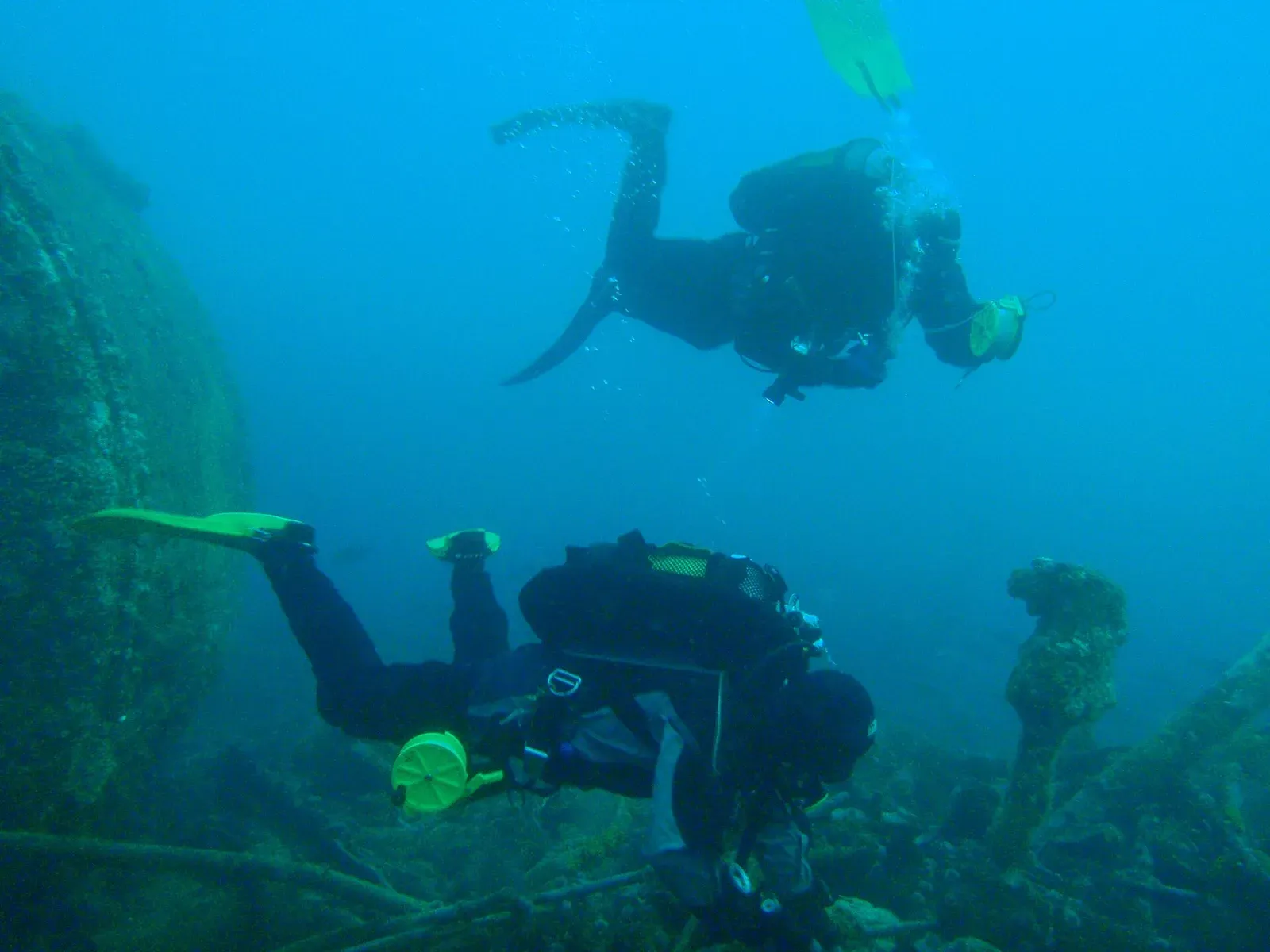 A diver above a wreck boiler holds a yellow reel and delayed surface marker buoy, with a second diver working below.