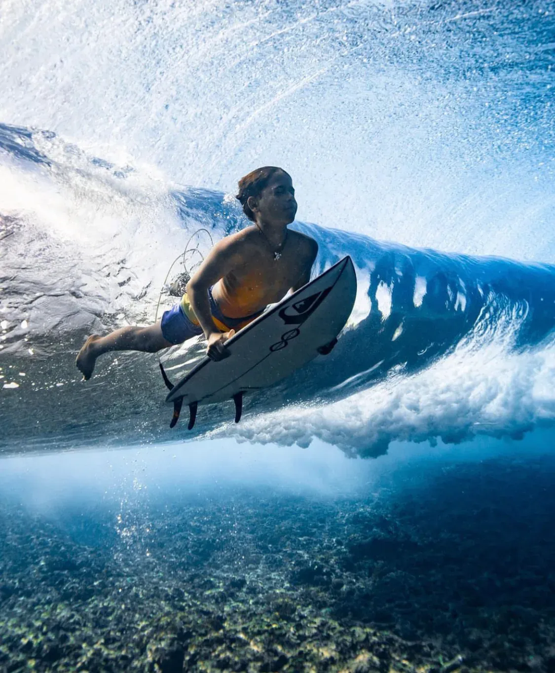 Seen from underwater, a surfer pushes his board down beneath the churning white underside of a breaking wave, with reef below.