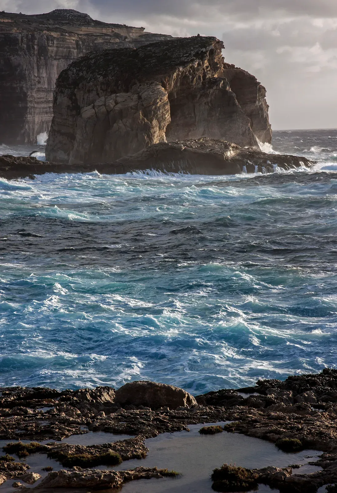 A rough sea heaves around Fungus Rock, a massive limestone islet at Dwejra on Gozo, under storm light.