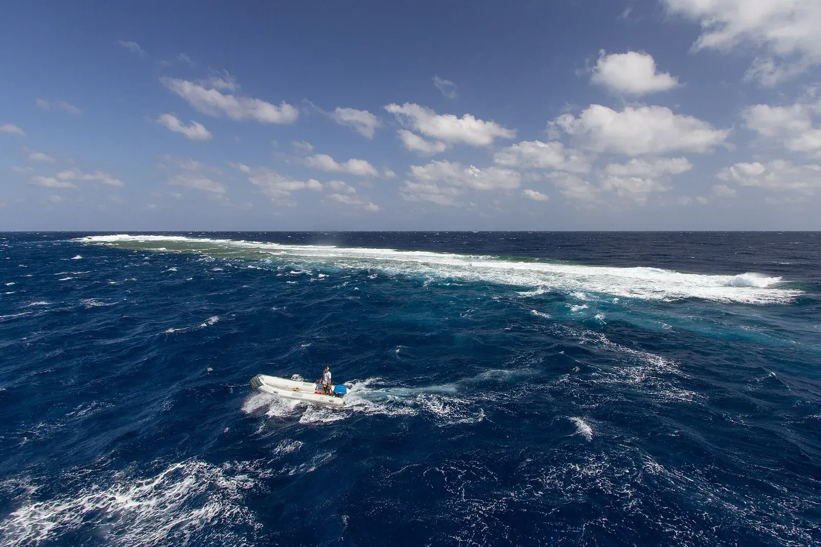 A small inflatable tender running across open blue ocean beside the white line of surf breaking over Elphinstone Reef, with no land in sight