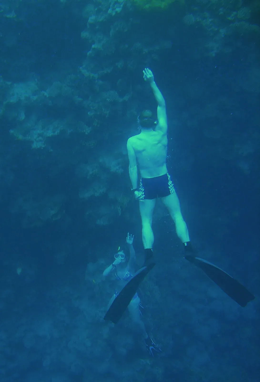A freediver in shorts and long fins hanging vertical, head up and fins down, by a dark reef wall, one arm raised overhead, a snorkeller below reaching up to him
