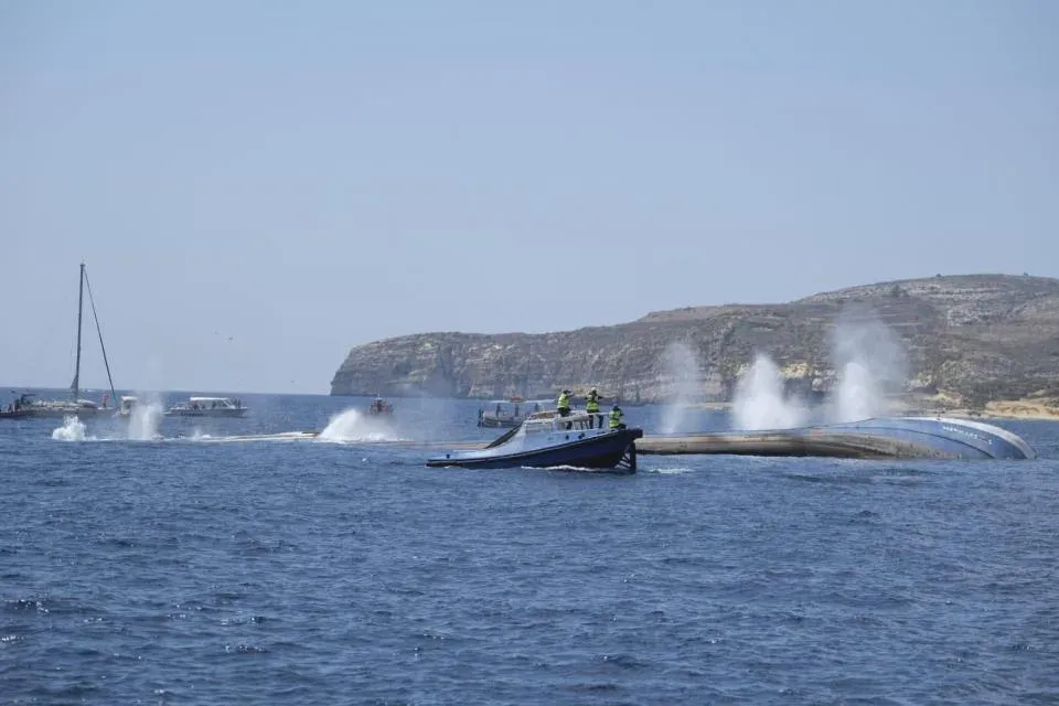 An old tanker's hull lying over on her side at the surface with water jets spraying, a work boat alongside and Gozo cliffs behind