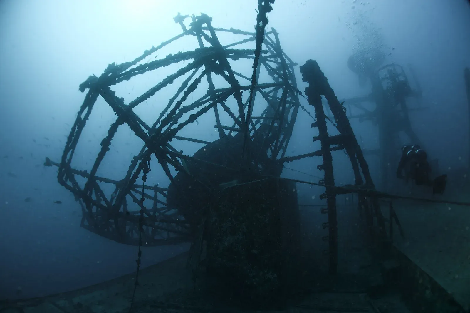 A diver in silhouette beside the encrusted lattice of a big radar dish on a sunken ship in blue water