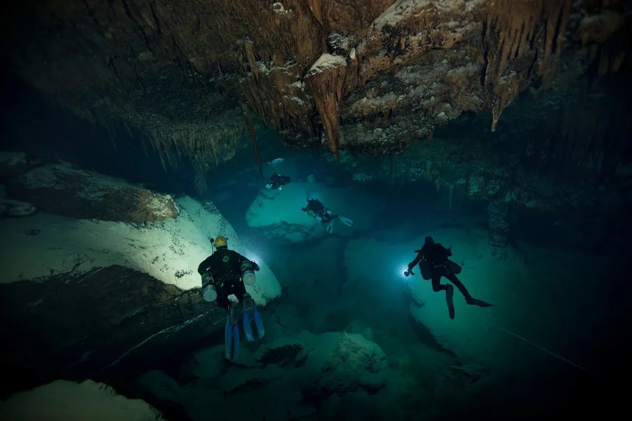 Four sidemount cave divers with lights crossing a large flooded chamber hung with stalactites, a guideline running across the floor