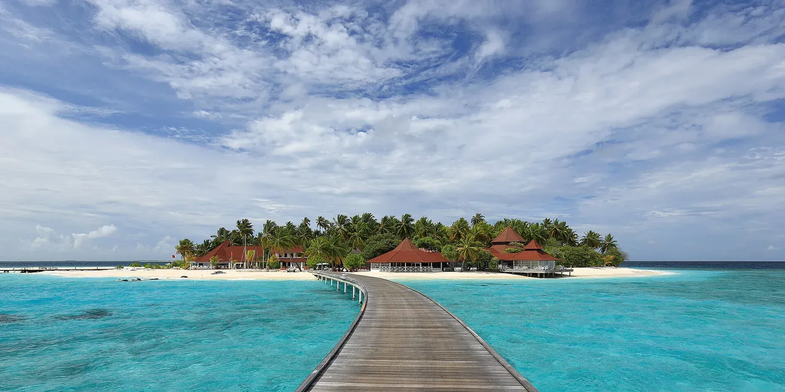 A long wooden jetty over a turquoise reef flat leading to a palm-covered resort island in the Maldives