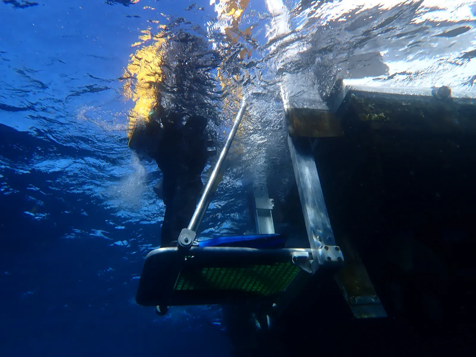 Seen from below the surface, a diver's dark silhouette climbs a boat's dive ladder into sunlight, with deep blue water all around.