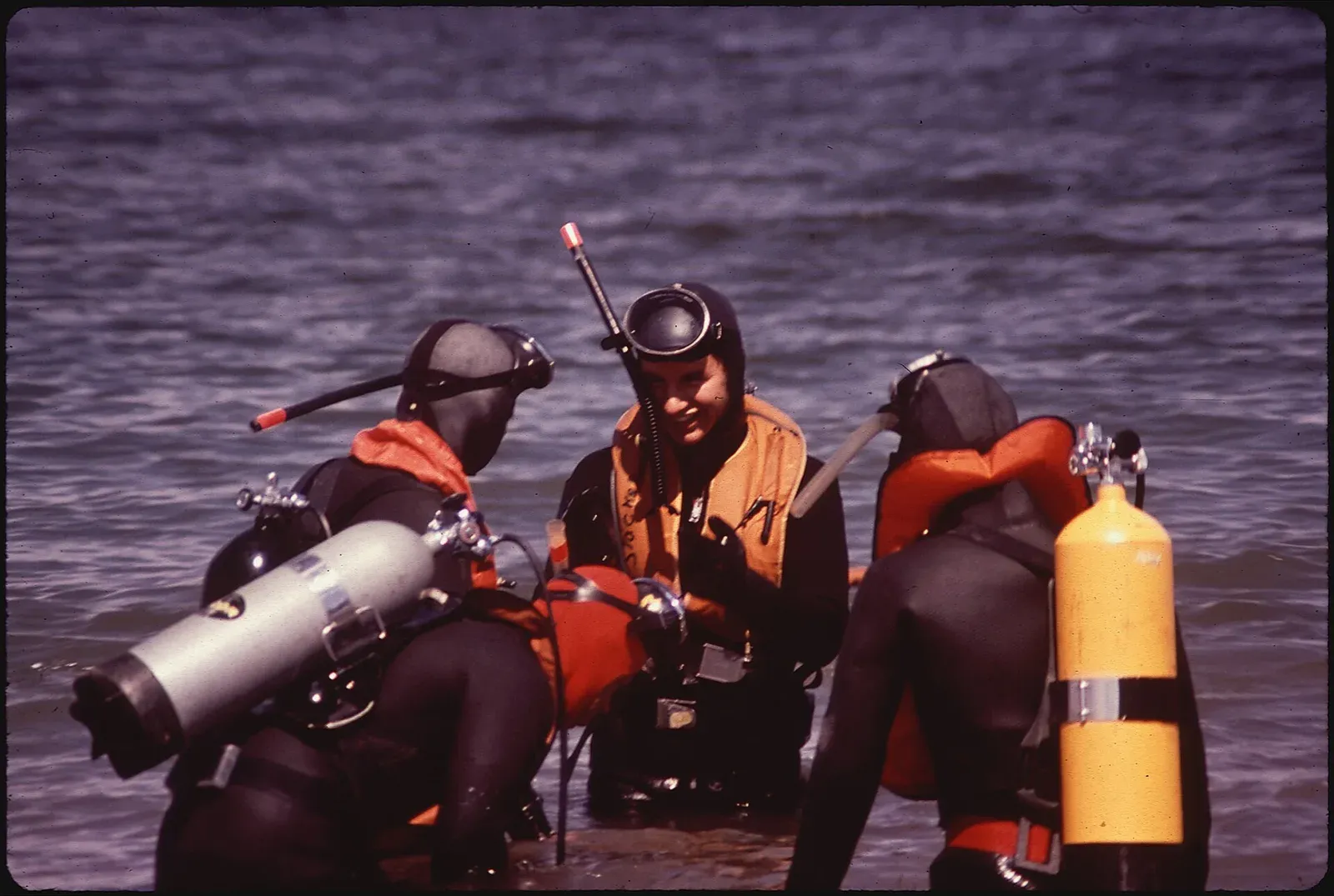 Three wetsuited divers with cylinders and orange flotation vests wade into the choppy blue water of Lake Powell in 1972.