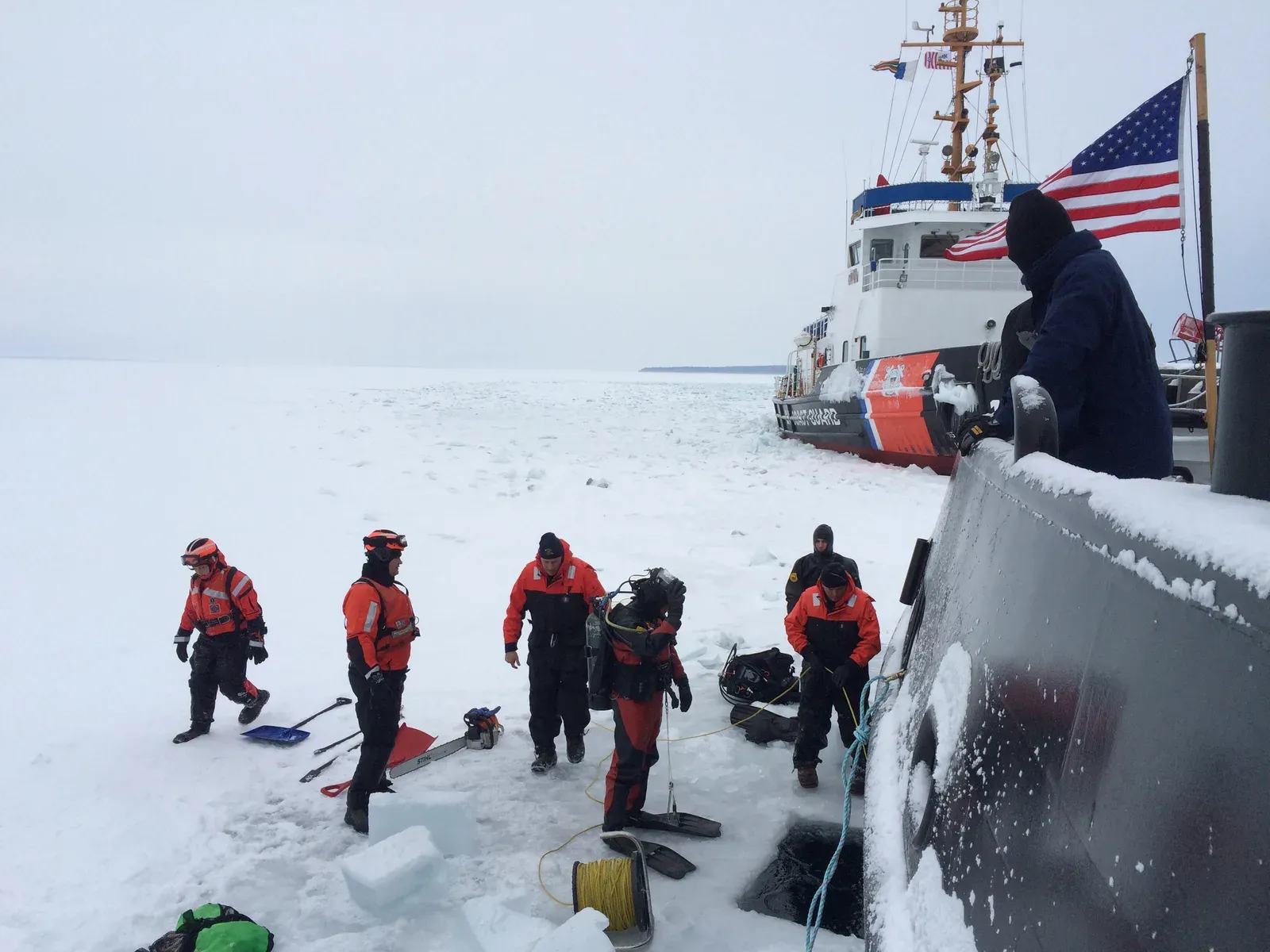 Drysuit divers on frozen water beside a Coast Guard cutter, with a chainsaw, lifted ice blocks and a reel of yellow line at the hole