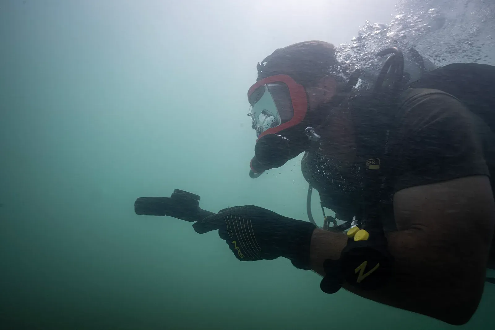 A diver in a full-face mask holds a compass out in front in bright, green, low-visibility water off Riviera Beach, Florida.