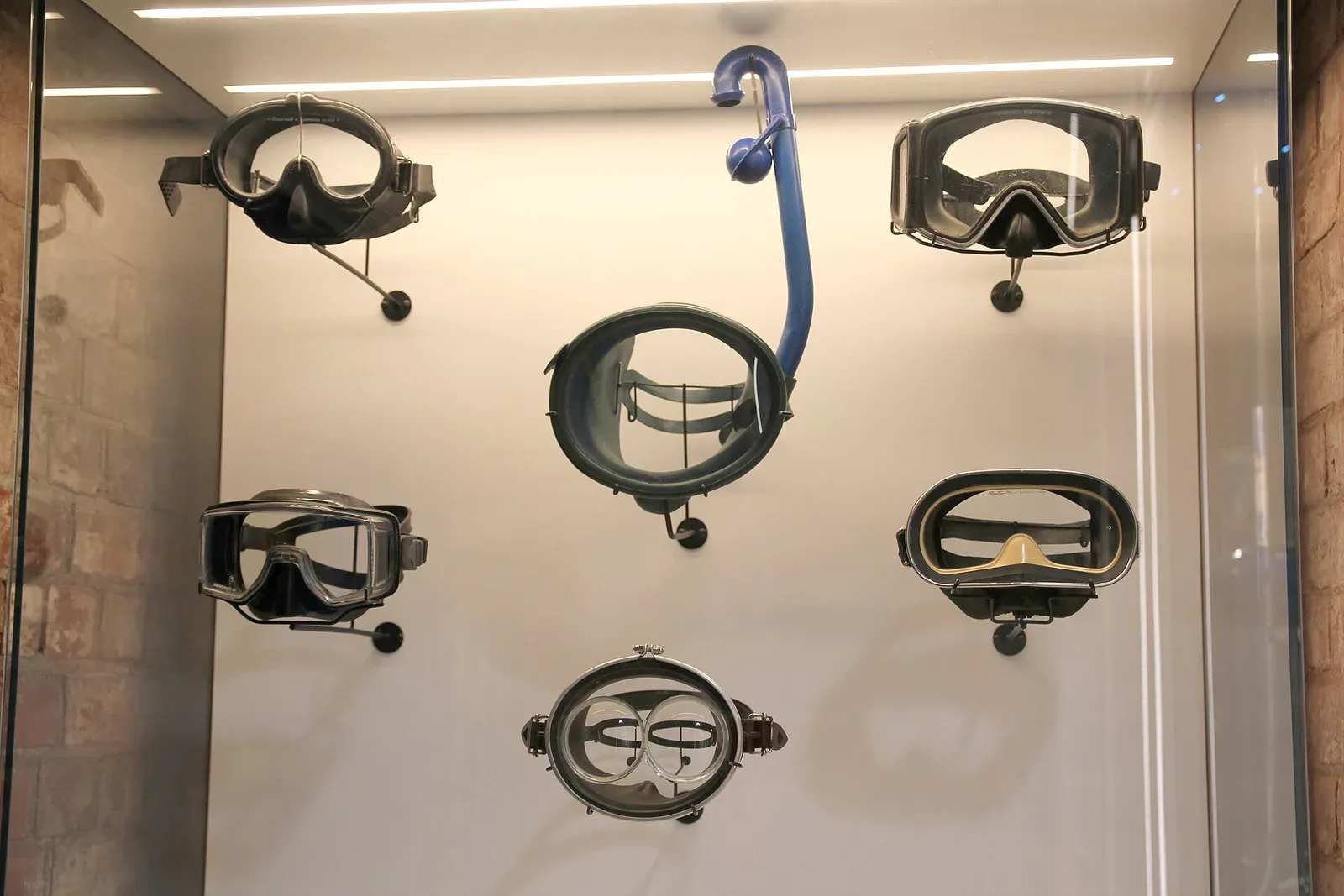Six diving and snorkelling masks of different eras mounted on a white panel in a museum case, one with a blue snorkel attached
