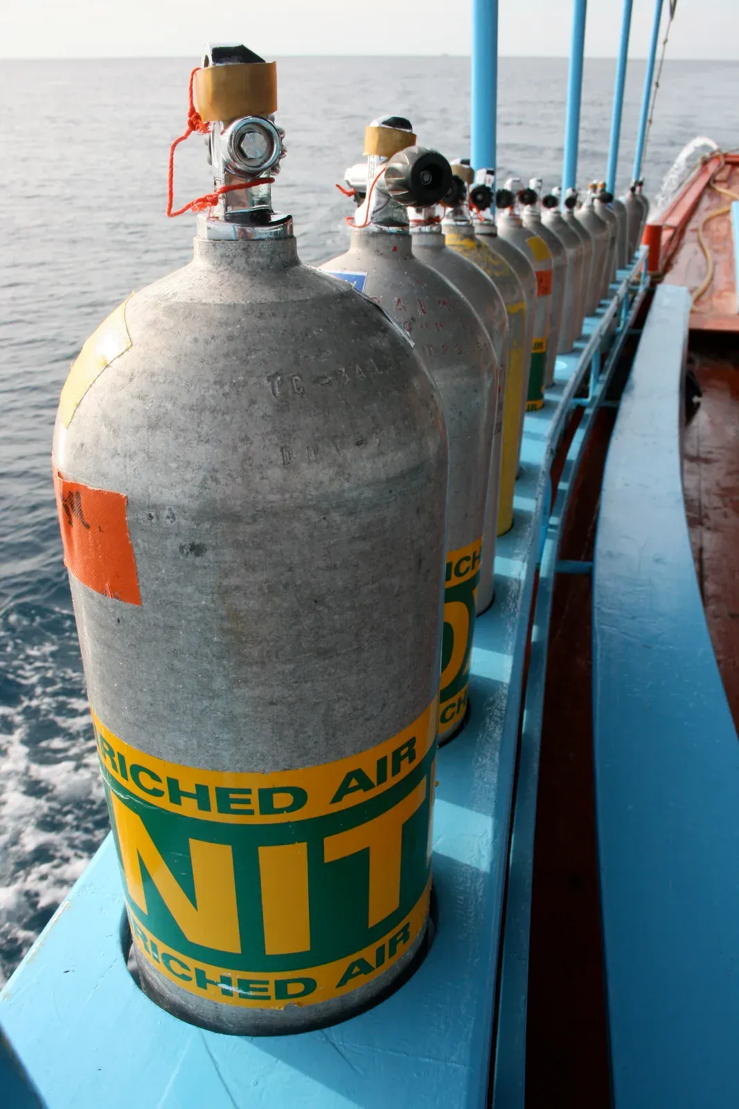 A rack of aluminium cylinders banded with green and yellow enriched air nitrox labels along a dive boat's rail
