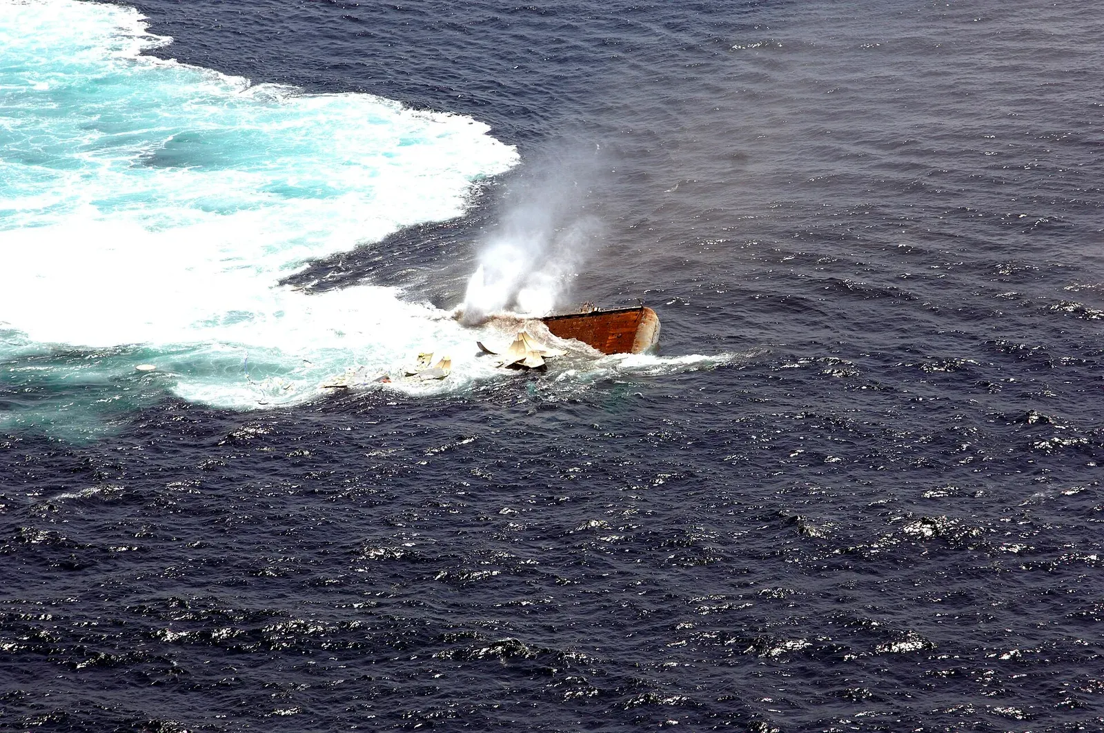 A rusty section of an aircraft carrier’s flight deck slipping under white water in open sea, air venting