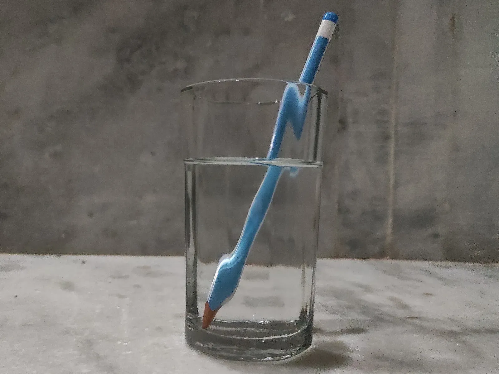 A blue pencil leaning in a glass of water, its submerged half appearing shifted and broken where it crosses the surface
