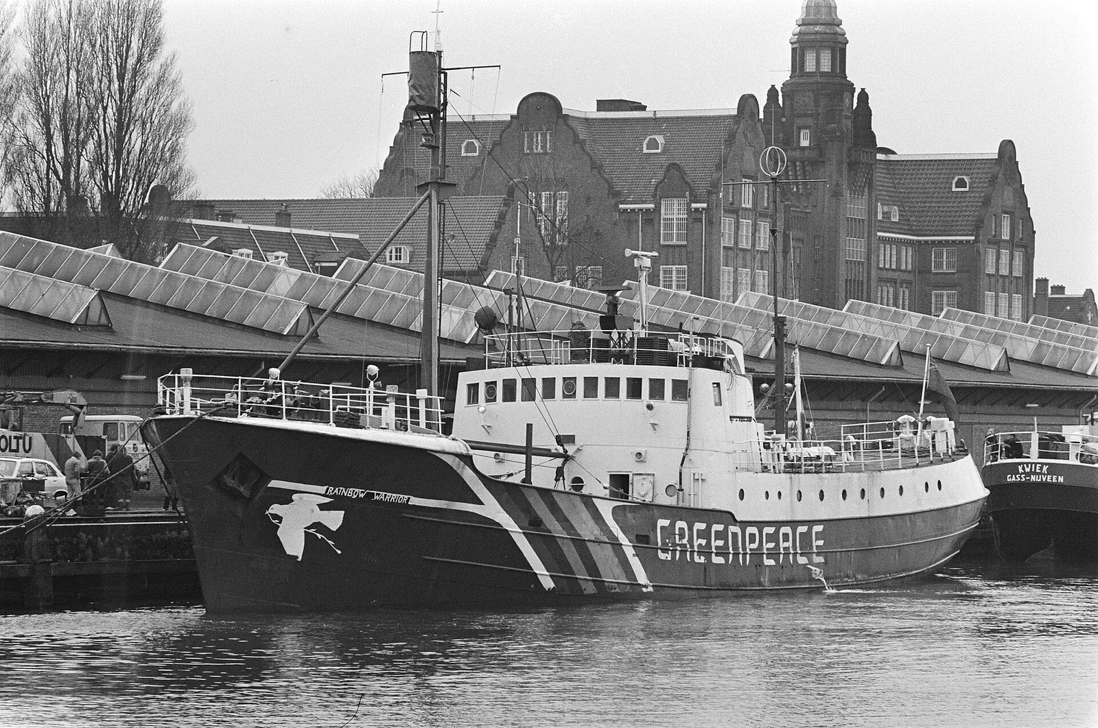 Black and white photo of a trawler painted with a dove and the word Greenpeace, moored in a city harbour