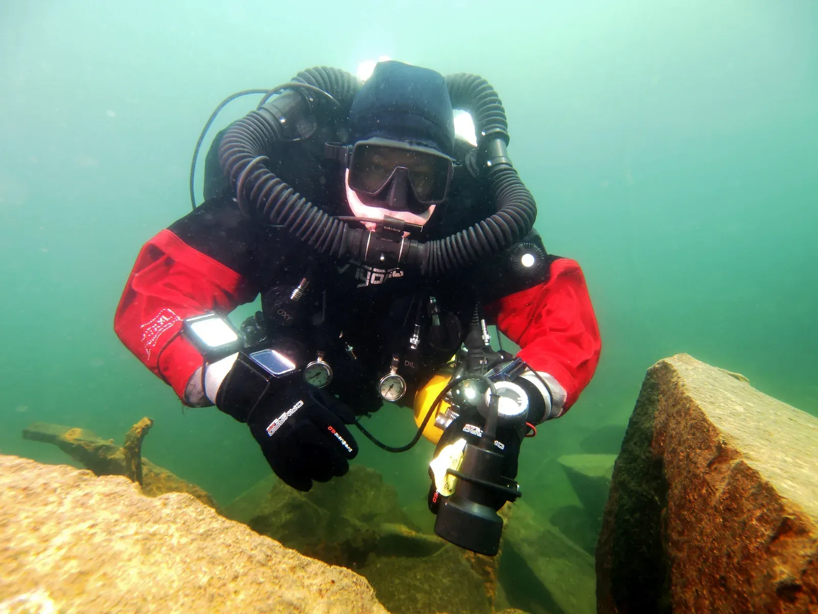 A diver on a closed-circuit rebreather with breathing hoses over both shoulders, hovering above a rocky bottom