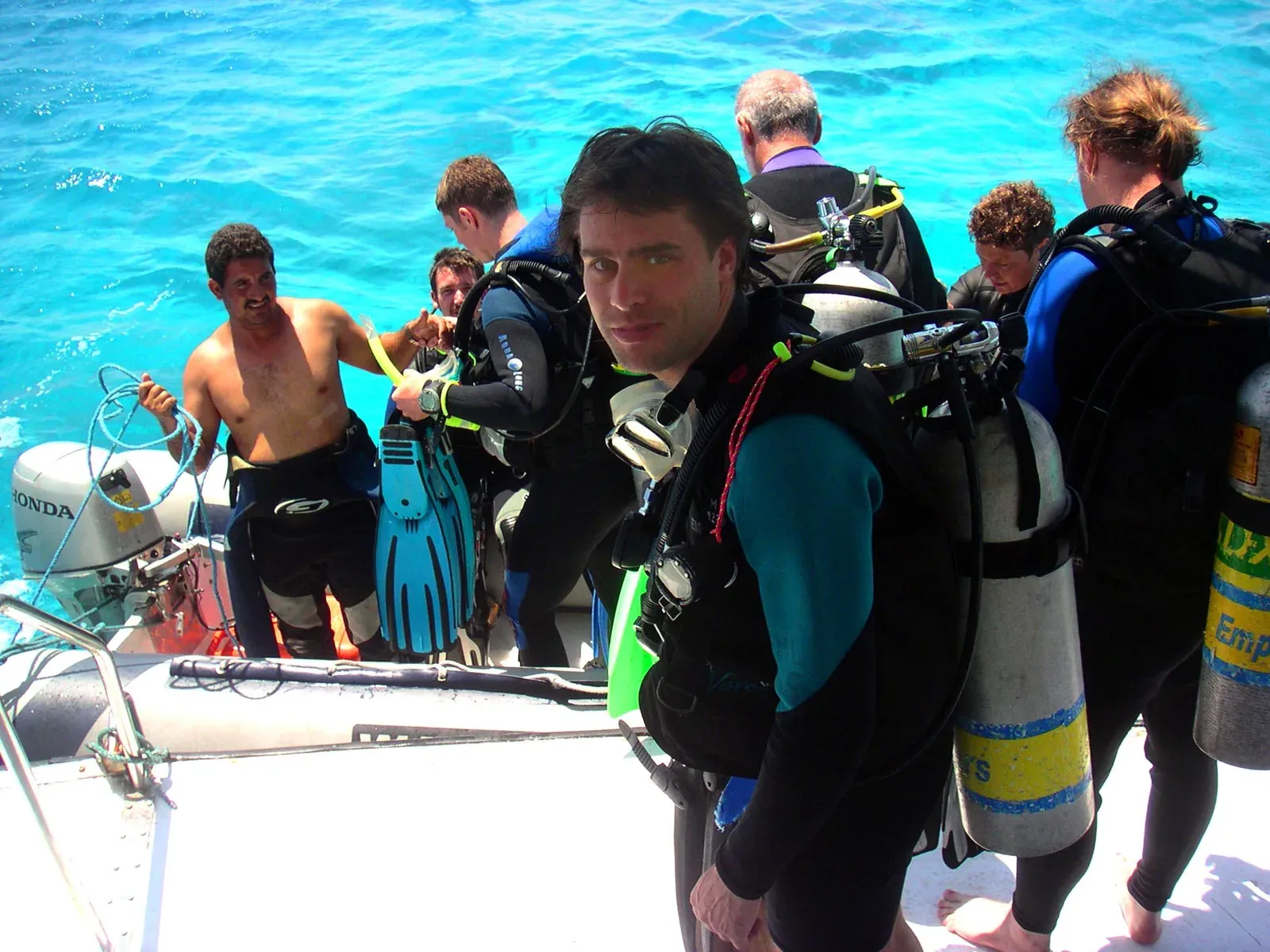 Divers in wetsuits with cylinders on their backs crowd the deck of a white dive boat beside a RIB with an outboard, turquoise sea behind.