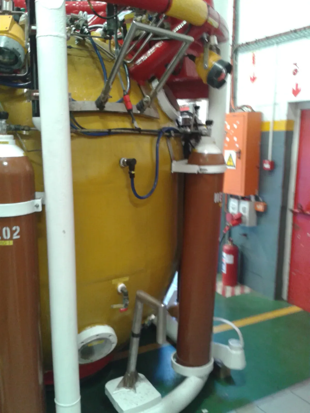 The yellow-painted exterior of a closed saturation diving bell in a workshop, with brown gas cylinders clamped to it, white pipework and a small round viewport