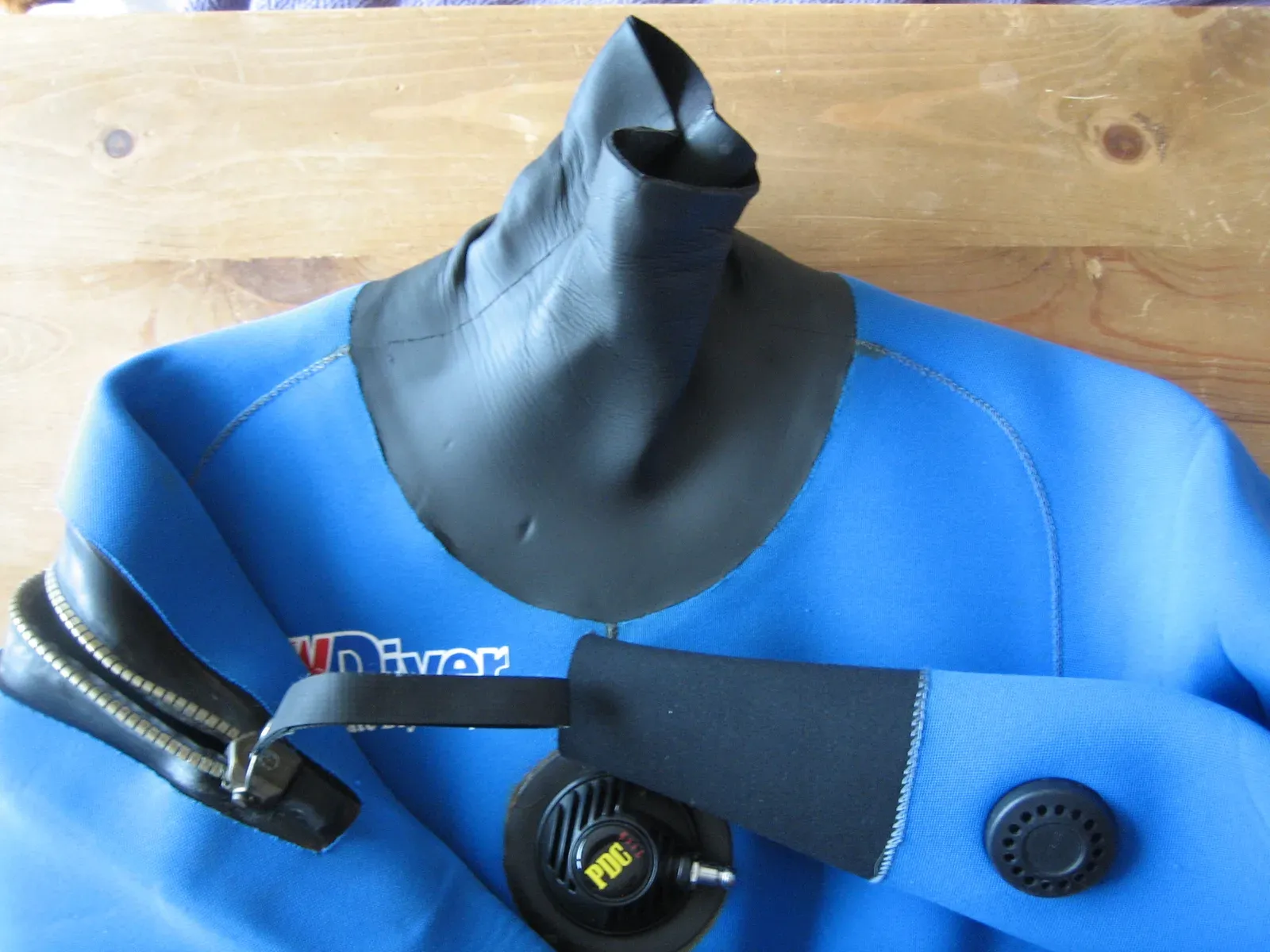 A blue neoprene drysuit laid on a wooden table, showing its black latex neck seal, a metal-toothed shoulder zip and a round valve on the chest.