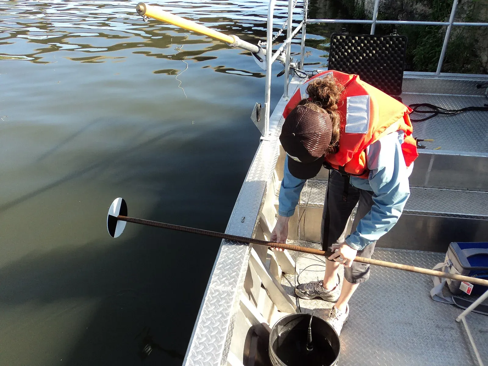 A researcher on a boat holds a black-and-white Secchi disk on a pole out over dark, murky water.