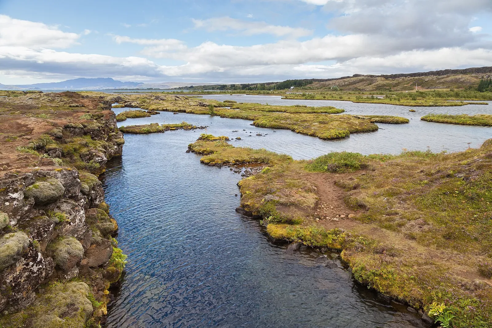 A narrow flooded rift of clear blue water cutting through moss-covered lava toward a lake and distant hills