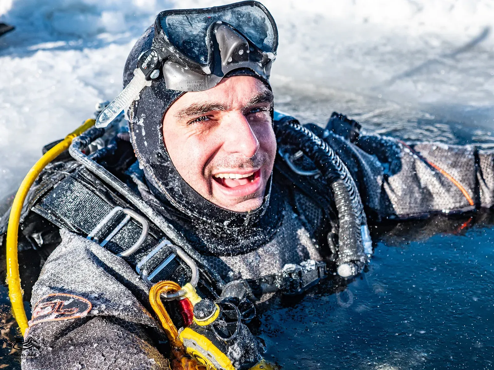 A drysuit diver surfacing in a hole cut through lake ice, frost forming on hood and regulator
