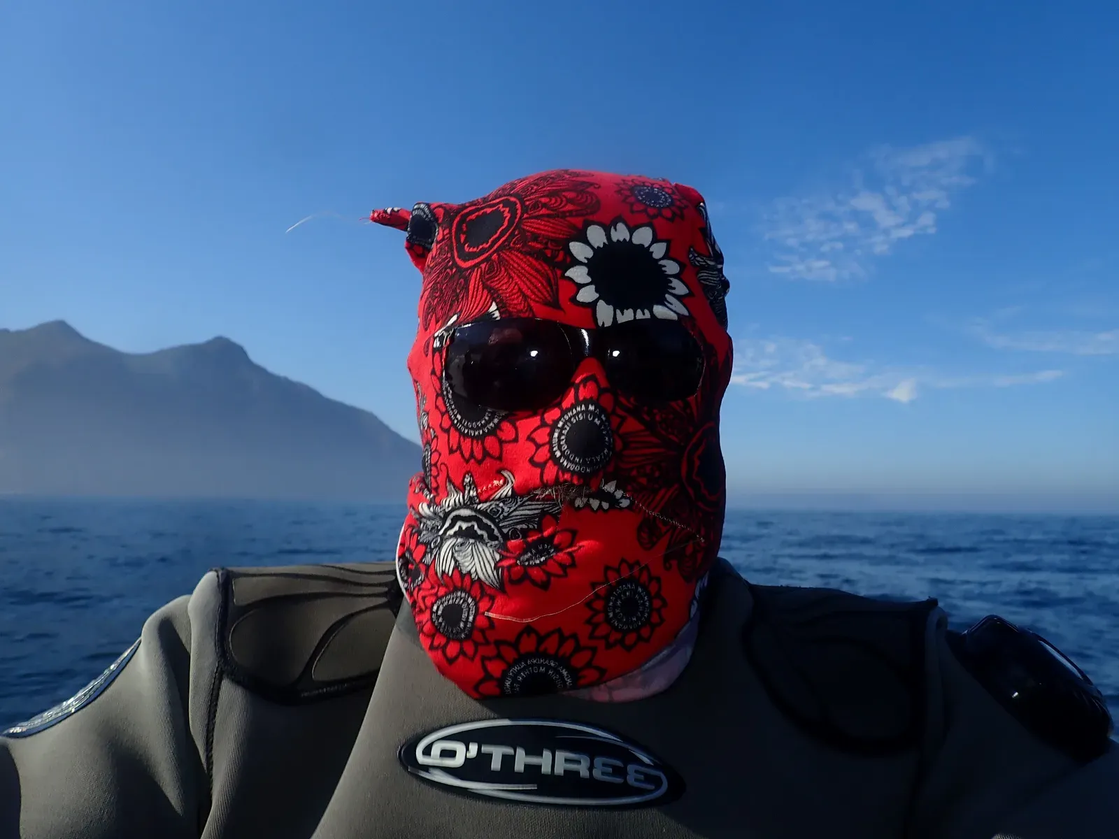 A diver in a grey exposure suit sits in a boat wearing a red floral full-face sun hood and dark sunglasses, with mountains and blue sea behind.