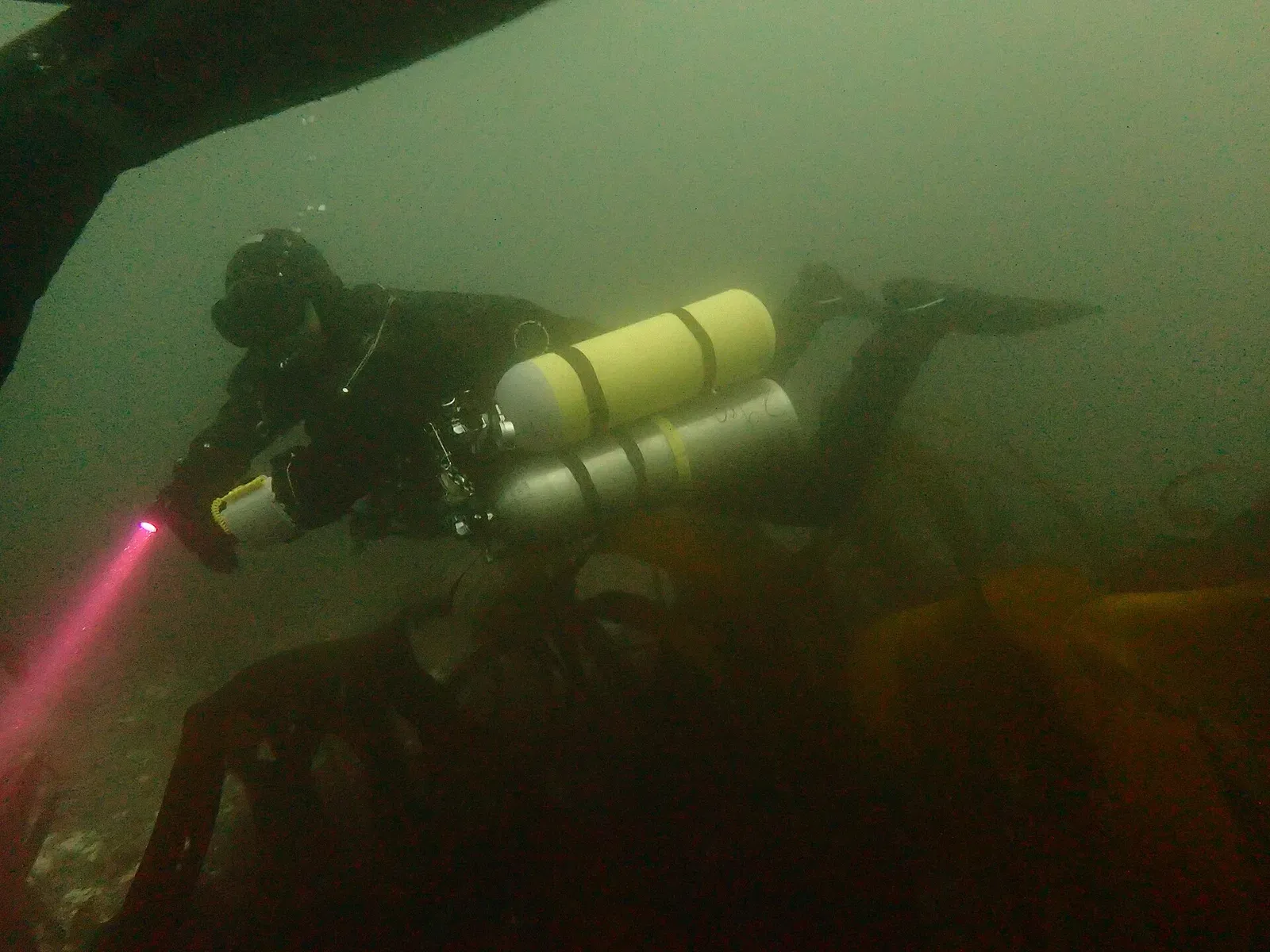 A drysuit diver carrying sidemount cylinders swims over a dim reef, a torch beam cutting through green water