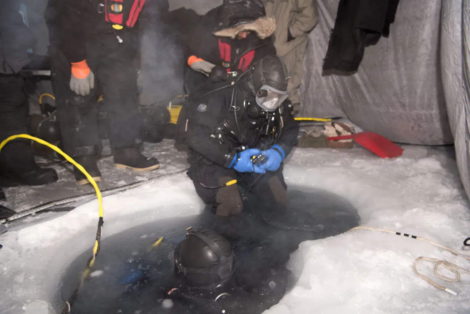 A diver in a full-face mask kneels at a hole cut through sea ice inside a tent, with lines laid on the ice beside the water
