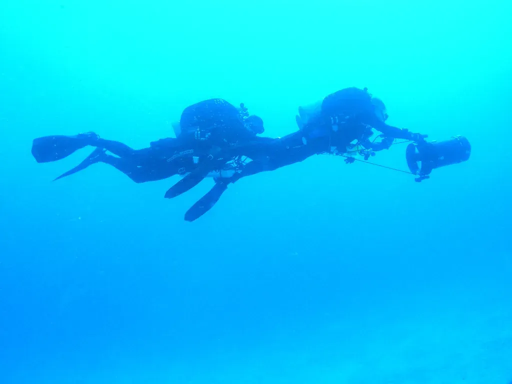 Two divers riding a single scooter in blue water, the second holding the ankle of the first