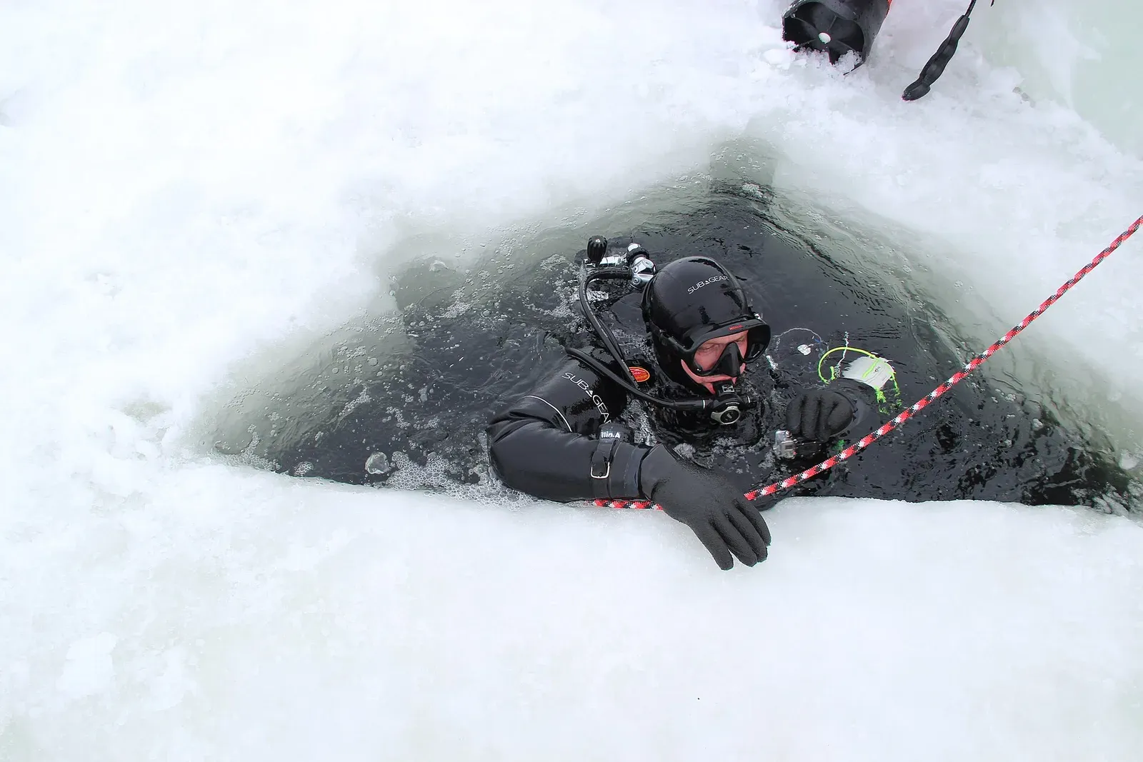 A diver in a black drysuit and hood at the surface of a triangular hole cut through thick ice, gripping a red-and-white line.
