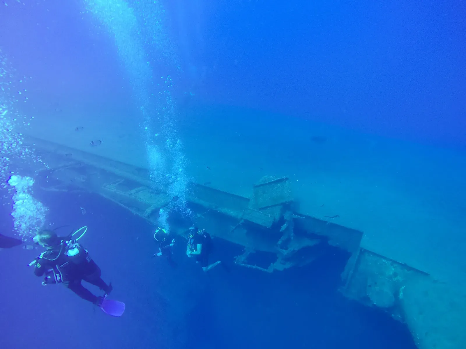 Three divers descending alongside the long sloping hull of a large shipwreck in clear blue water